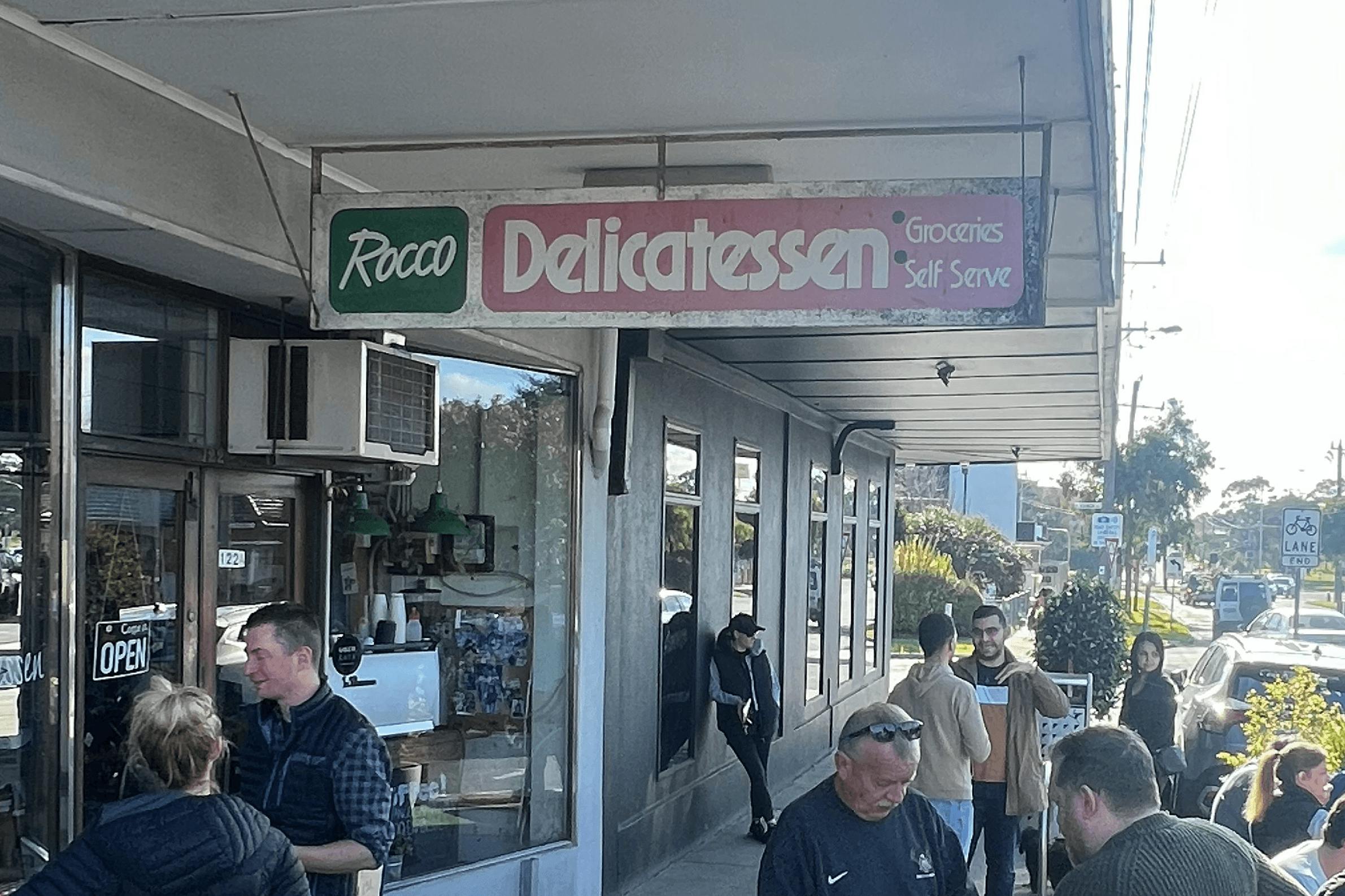 The outdoor of a deli with people standing around.